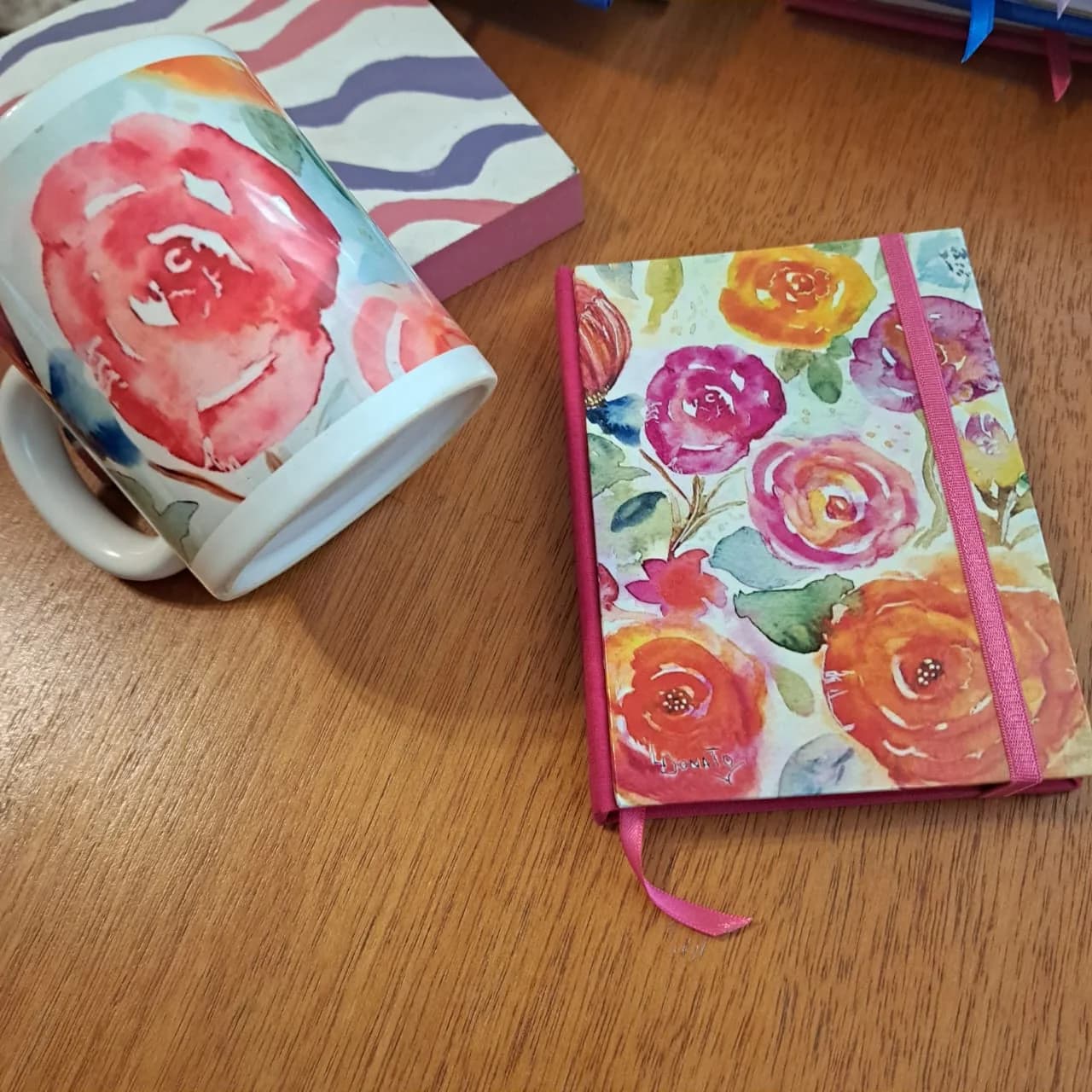 Taza y cuaderno de tapa dura con acuarelas de rosas en rojos, naranjas y fucsias.